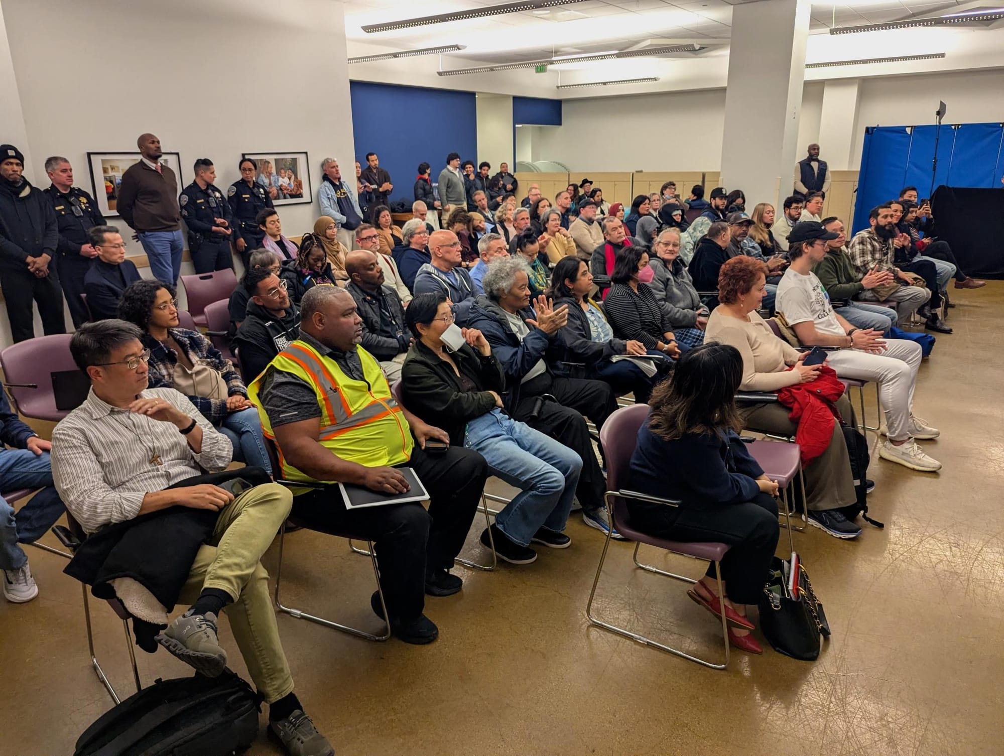 People sit in four rows of chairs arranged in a community room, with more  standing against a back wall.